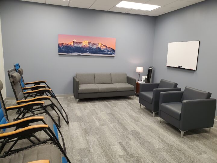 Group therapy room with couch and zero gravity chairs. There is a painting on the wall of mountains and a whiteboard. 