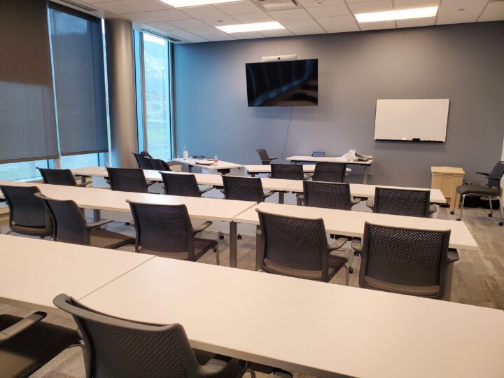 Conference room with 3 rows of long tables with black chairs. A large TV and whiteboard are at the front of the room. 