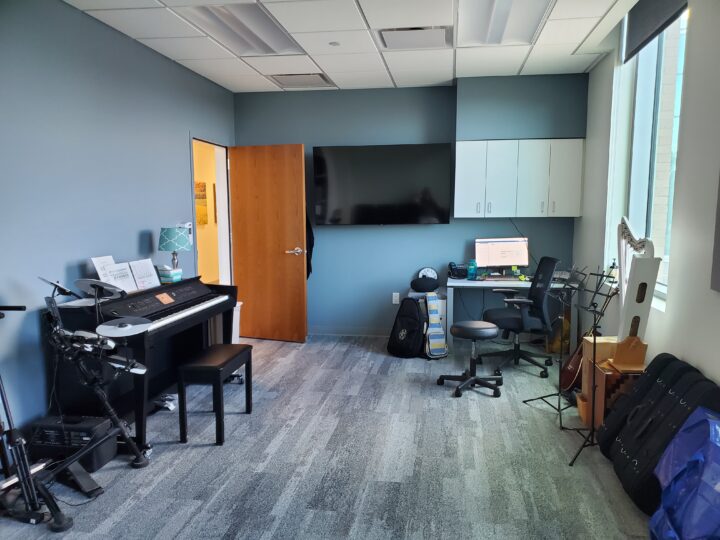 Music therapy room including a piano with a piano bench and cymbals. White cabinets and TV are mounted on the wall. In the corner is an office chair with a desk and computer. There is a row musical instrument cases along the wall and sheet music stands.