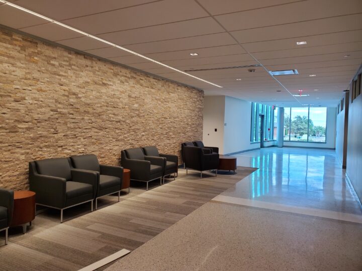 Hallway with comfortable chairs and end tables along a stone wall. Big windows at end of hallway. 